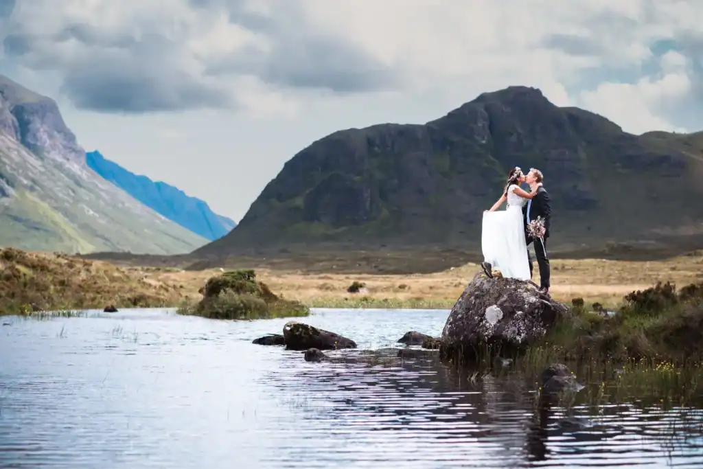 Bride and groom embracing on scenic rock by a lake on the Isle of Skye with a mountain backdrop