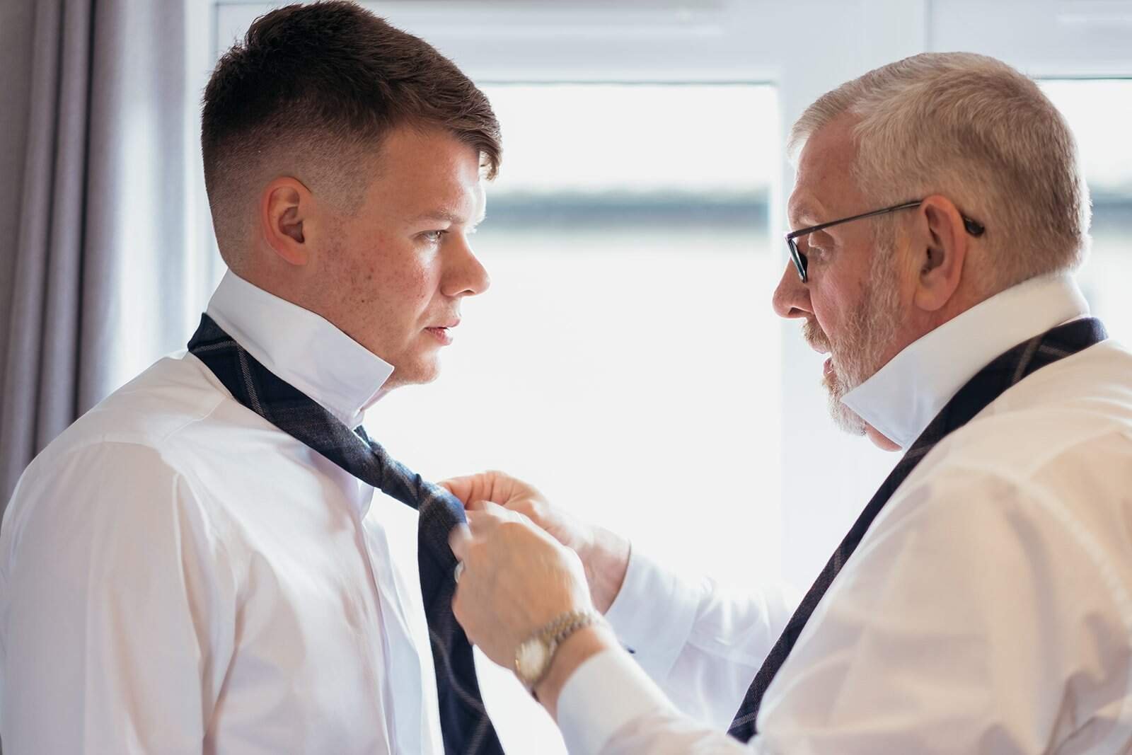 Elderly man helping young man with necktie near window