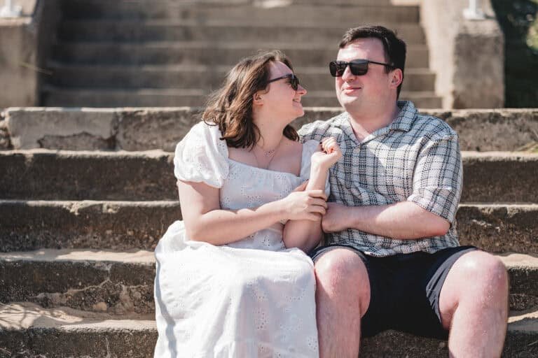 A smiling couple sitting on stone steps in summertime at Seaburn for their engagement photoshoot