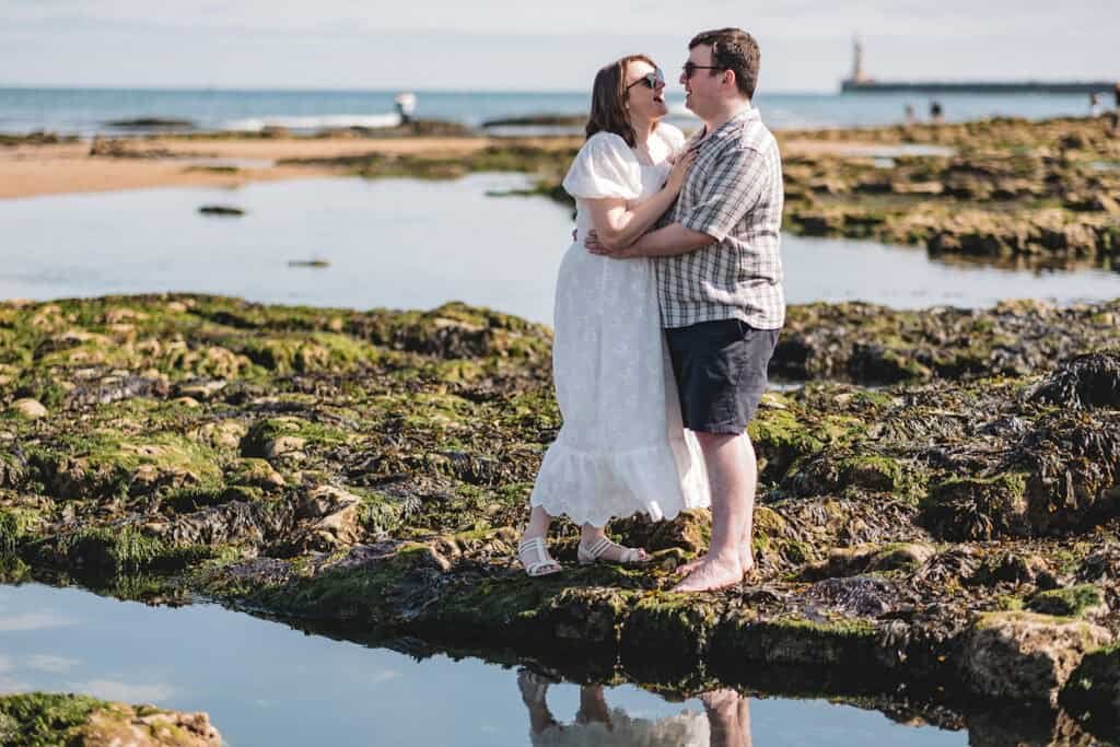 Couple embracing on rocky beach with lighthouse in background