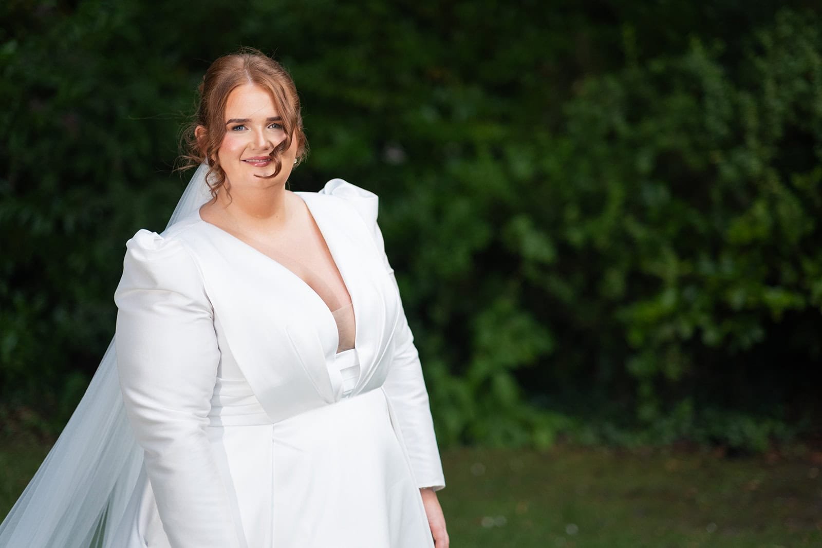Bride in elegant white dress smiling outdoors against greenery