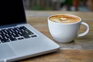Close-up of a latte coffee with heart art next to a laptop on a rustic wooden table.