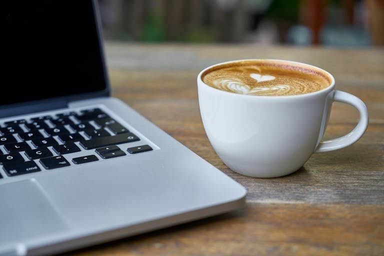 Close-up of a latte coffee with heart art next to a laptop on a rustic wooden table.